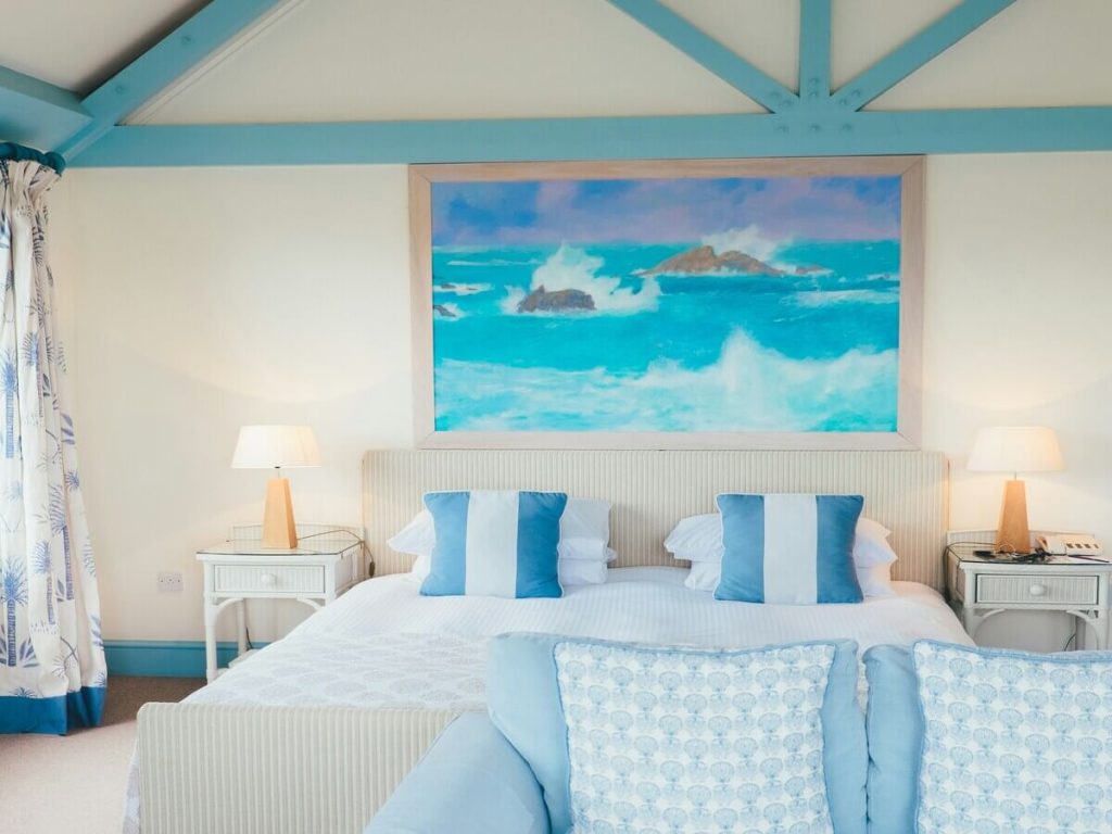 A serene bedroom with soft blue and white decor, featuring a large coastal-themed painting above the bed, blue beams on the ceiling, and natural light streaming in, creating a calm and airy atmosphere