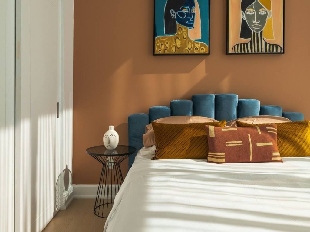 A stylish bedroom with a warm brown accent wall, contemporary art, a blue upholstered headboard, and decorative pillows in earthy tones, bathed in soft, natural sunlight for a cozy, artistic look