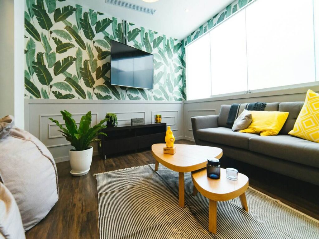 A modern living room with a bold tropical leaf wallpaper, a gray sofa with yellow accent pillows, a wooden coffee table, and natural light pouring through large windows, giving the space a fresh, vibrant feel