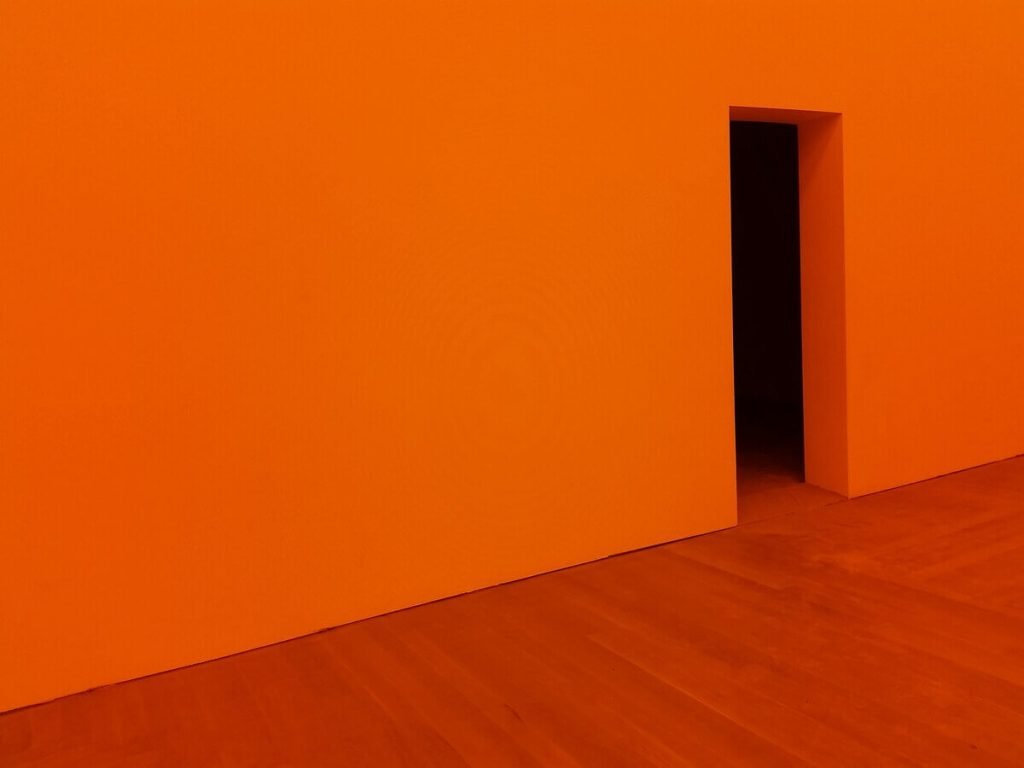 A minimalist room with a vivid orange wall and an open doorway, featuring a simple wooden floor, creating a bold and striking nature color palettes with an abstract, modern feel