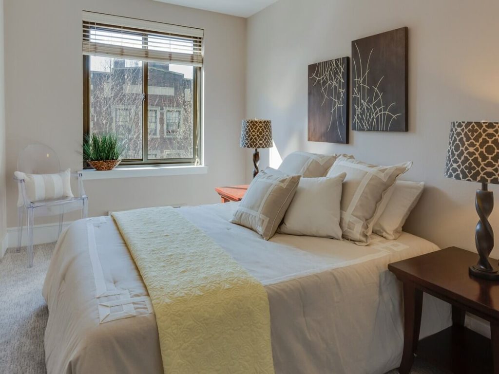 A cozy and contemporary bedroom with neutral tones, featuring a large window, soft bedding, a wooden nightstand with a lamp, and modern abstract wall art, creating a warm and inviting space