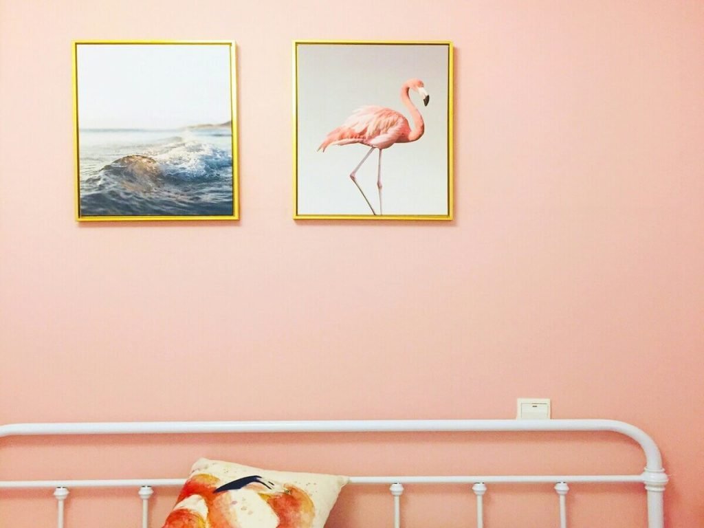 A minimalist bedroom with pastel pink walls, featuring framed artwork of a wave and a flamingo, a simple white metal bed frame, and a matching flamingo pillow, creating a soft and playful aesthetic