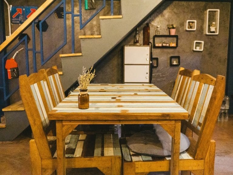 A rustic dining setup featuring a wooden table with a striped design and matching chairs, set against an industrial-style staircase with blue metal railings and a textured gray wall.